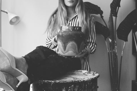 a black and white image of a girl sitting with her legs crossed over a muddied pottery wheel. she is holding a large vase in her lap.